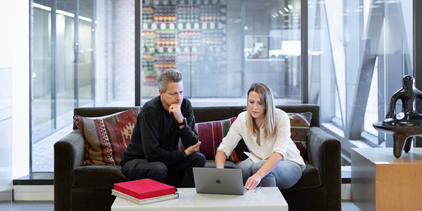 Man and women looking at laptop on sofa with glass office behind