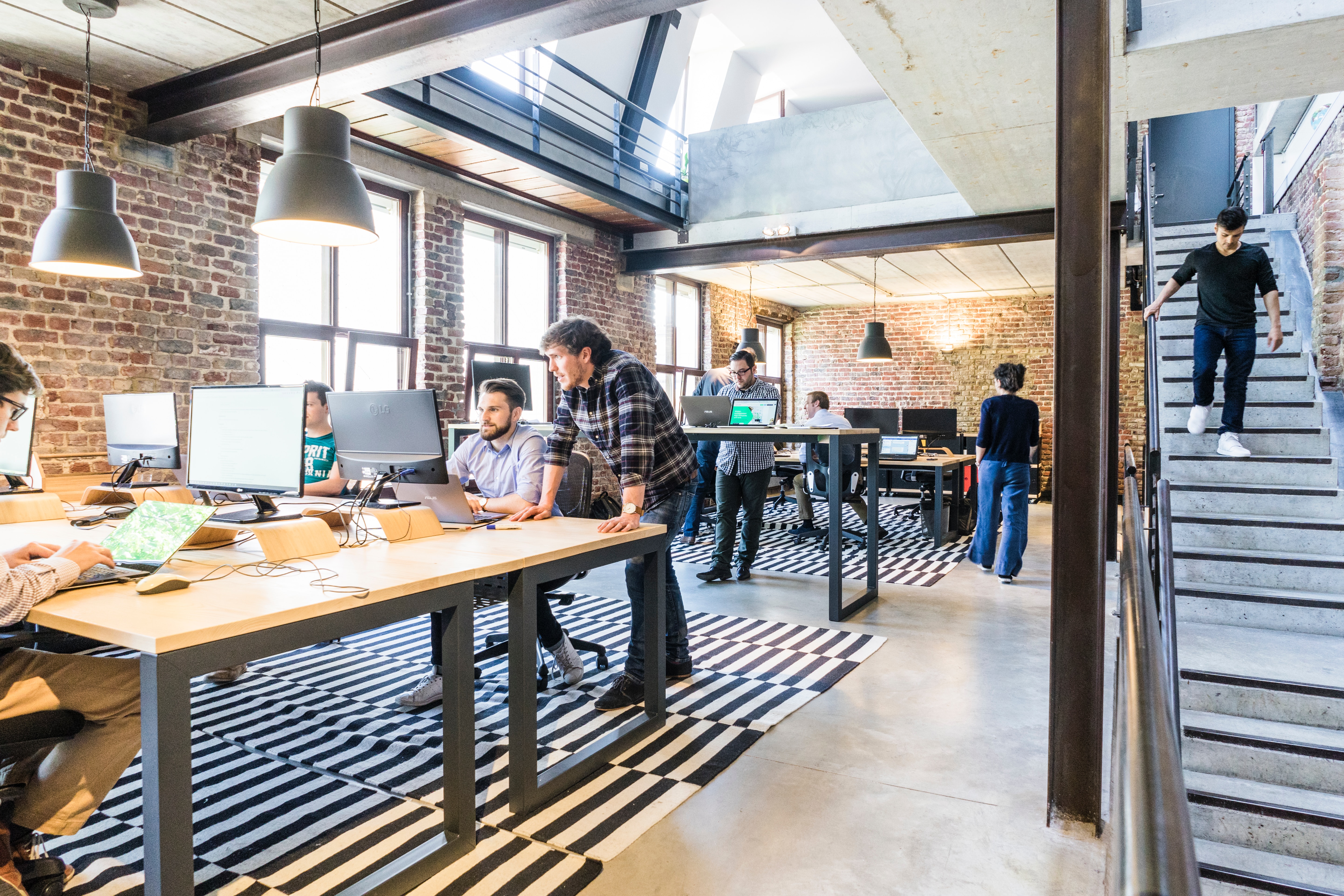 Open plan office with long tables and laptops with brick interior and wooden floor and stairs